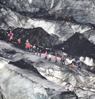 A group of people equipped with climbing gear, including helmets and backpacks, is traversing a rugged and icy glacier landscape. The terrain features sharp contrasts between the dark, rocky surfaces and lighter icy patches.