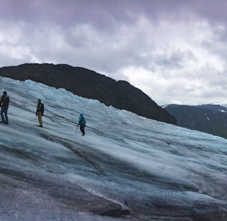A group of people are trekking across a vast, icy glacier with snow-capped mountains in the background under a cloudy sky. They are connected by a rope for safety, wearing snow gear and carrying climbing equipment. The scene conveys a sense of adventure and exploration amid a rugged natural landscape.