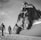 Two climbers are trekking across a snow-covered landscape towards a towering, rugged mountain peak. They are equipped with climbing gear and appear determined as they progress through the icy environment. The scene is monochromatic, giving it a timeless and adventurous feel.
