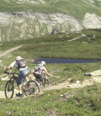 Two cyclists are navigating a rugged mountain trail. The landscape features lush green hills, a rocky path, and a small body of water in the distance. Both cyclists are wearing helmets and backpacks, emphasizing the outdoor adventure theme.