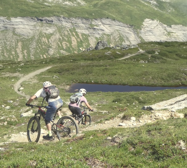 Two cyclists are navigating a rugged mountain trail. The landscape features lush green hills, a rocky path, and a small body of water in the distance. Both cyclists are wearing helmets and backpacks, emphasizing the outdoor adventure theme.