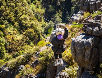 A person is zip-lining across a lush, green landscape, surrounded by dense foliage and rocky outcrops. The scene depicts adventure and nature, with the individual wearing a helmet and harness for safety.