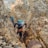 A group of climbers in rugged outdoor gear navigate a narrow, rocky crevice on a mountain. The scene is set amid large, jagged boulders, with some snow-capped peaks visible in the background. Each climber wears helmets and backpacks, emphasizing the challenging and adventurous nature of their ascent.