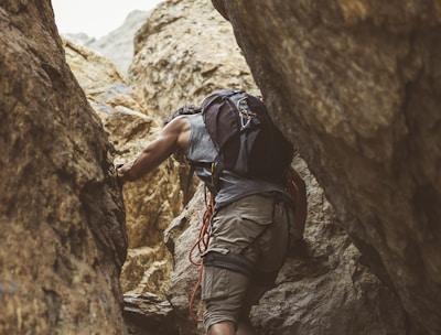 A person wearing casual climbing attire navigates through a rugged, narrow rock crevice. They are equipped with a backpack and climbing gear, including a rope looped around their shoulder. The scene captures a sense of adventure and challenge against a backdrop of textured, earthy rock formations.