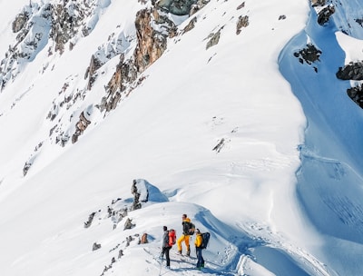 A group of four people wearing colorful winter clothing engage in a mountaineering or skiing adventure on a snow-covered mountain slope. The slope is steep, with visible tracks in the snow indicating their path. Jagged rocks protrude through the snow, adding to the rugged terrain.