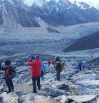 Several hikers are trekking along a rocky, mountainous path surrounded by snow-capped peaks. The landscape is rugged and expansive, with layers of distant mountains. The group wears colorful hiking gear, including jackets and backpacks.