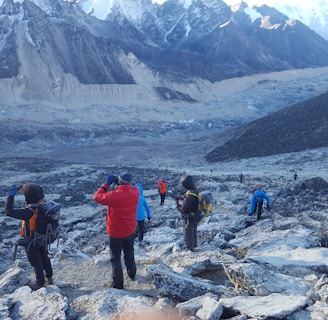 Several hikers are trekking along a rocky, mountainous path surrounded by snow-capped peaks. The landscape is rugged and expansive, with layers of distant mountains. The group wears colorful hiking gear, including jackets and backpacks.