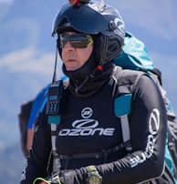 A person is wearing a helmet with a camera mounted on top, sunglasses, and a jacket labeled with 'Ozone'. They are equipped with various gear including a radio and a wristwatch, and appear ready for an outdoor adventure, possibly paragliding. The background features a blurred mountainous landscape.