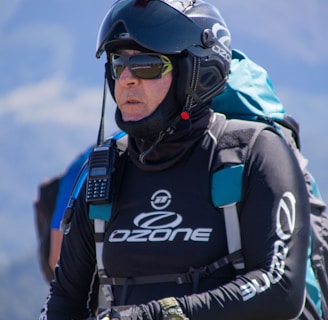 A person is wearing a helmet with a camera mounted on top, sunglasses, and a jacket labeled with 'Ozone'. They are equipped with various gear including a radio and a wristwatch, and appear ready for an outdoor adventure, possibly paragliding. The background features a blurred mountainous landscape.
