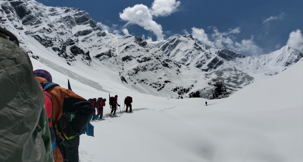 A group of hikers is trekking through a snowy mountainous landscape under a clear blue sky. The snow-covered mountains loom majestically in the background, while the hikers wear colorful gear and carry backpacks. The scene suggests a challenging and adventurous journey through pristine, untouched nature.